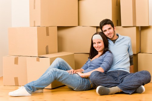 A young couple is seated on a hardwood floor inside a residential property, smiling at the camera while surrounded by numerous large cardboard moving boxes. The woman, wearing a white headband, a blue long-sleeved top, and light blue jeans, is leaning back against the man with her legs extended out. The man, dressed in a light blue shirt and dark blue jeans, is seated behind her, with his arms resting around her and both appear relaxed. The boxes are stacked behind them, some positioned directly against the wall, indicating a moving or packing activity within a home in Merton. The environment suggests an indoor residential space with natural lighting, and the scene visually highlights aspects of house removals, packing, and household belongings typical of local moving services in the Merton area.
