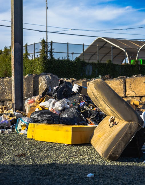 A large green waste disposal dumpster filled with assorted cardboard boxes, including flattened and partially assembled cartons, and packaging materials such as plastic wrap and paper padding, positio
