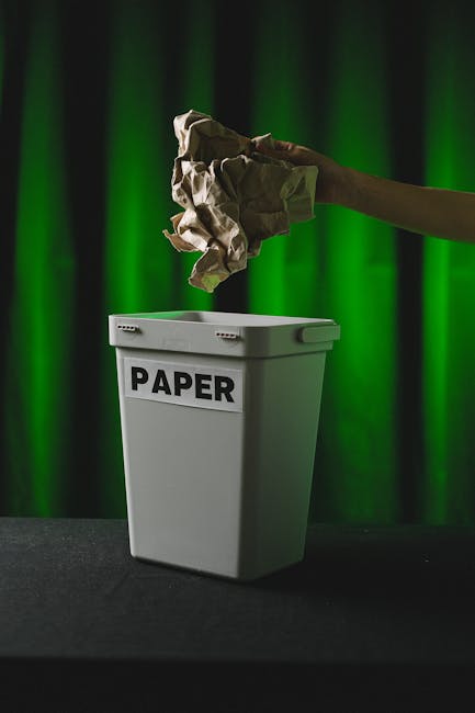 A person's arm is extending into the frame from the right side, holding a crumpled piece of brown paper above a white paper recycling bin labeled 'PAPER'. The bin is positioned on a dark surface with 