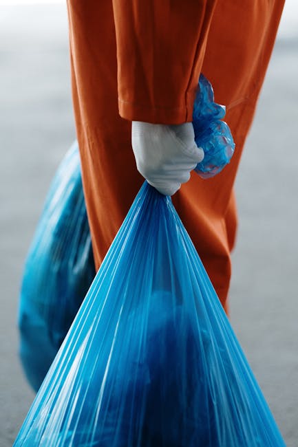 A person's arm is extending into the frame from the right side, holding a crumpled piece of brown paper above a white paper recycling bin labeled 'PAPER'. The bin is positioned on a dark surface with 